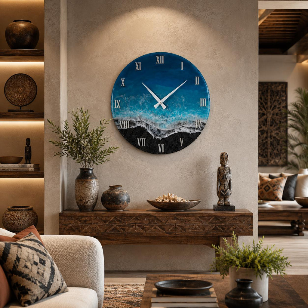 A circular wall clock featuring a resin art design that mimics an ocean shoreline with deep blue water, turquoise waves, white sea foam, and a black sand beach. The clock face displays white Roman numerals and two white clock hands.