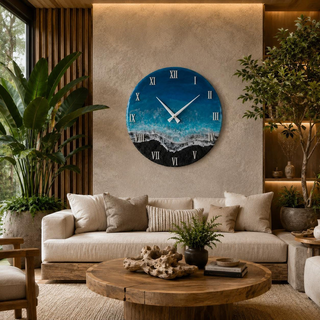 A circular wall clock featuring a resin art design that mimics an ocean shoreline with deep blue water, turquoise waves, white sea foam, and a black sandy beach. The clock face displays white Roman numerals and two thin white hands.