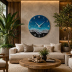 A circular wall clock featuring a resin art design that mimics an ocean shoreline with deep blue water, turquoise waves, white sea foam, and a black sandy beach. The clock face displays white Roman numerals and two thin white hands.