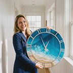 A large circular wall clock featuring a vibrant ocean wave design with turquoise water, white sea foam, and a sandy beach shore. The clock has a silver-toned metal frame with classic Roman numerals and two thin silver clock hands.