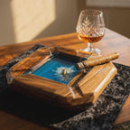 A square wooden ashtray with a central blue resin inlay featuring a small gold-colored whale figurine amidst white foam details. A brown cigar rests across the rim of the ashtray, positioned near a crystal glass filled with amber liquid.