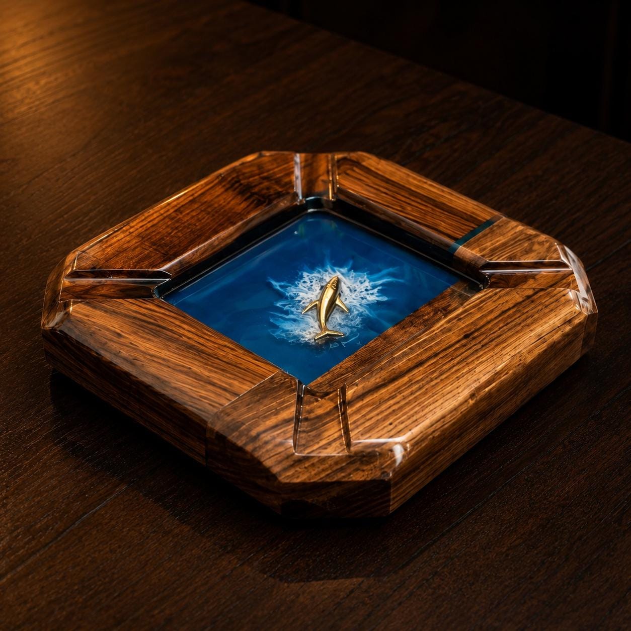 A square wooden ashtray featuring a polished dark brown wood grain frame with four notches. The center contains a deep blue resin inlay depicting a swirling ocean wave with a small, metallic gold-colored shark figure in the middle.