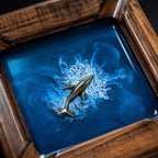 A square wooden tray featuring a deep blue resin inlay that mimics ocean water. A metallic gold-toned whale figurine is embedded in the center, surrounded by white swirling patterns that resemble sea foam and crashing waves.