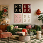 Four square wall art panels arranged in a grid, each featuring the word KISS in large cut-out letters. The top panels are black and dark red, while the bottom panels are light pink and white, all displayed above a green velvet sofa.